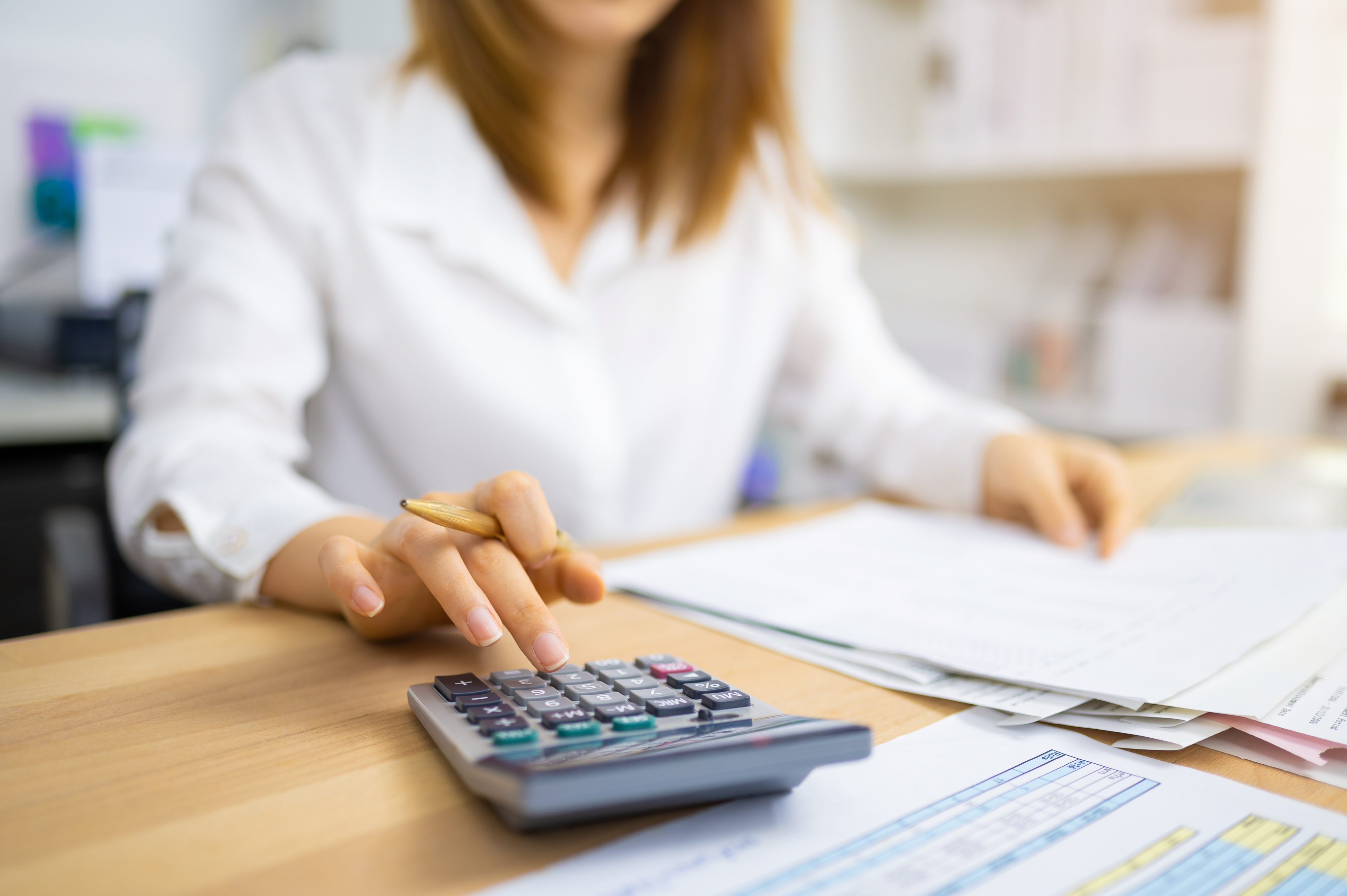 A person working at a desk is using a calculator while holding papers. The image has a professional setting with documents spread on the desk