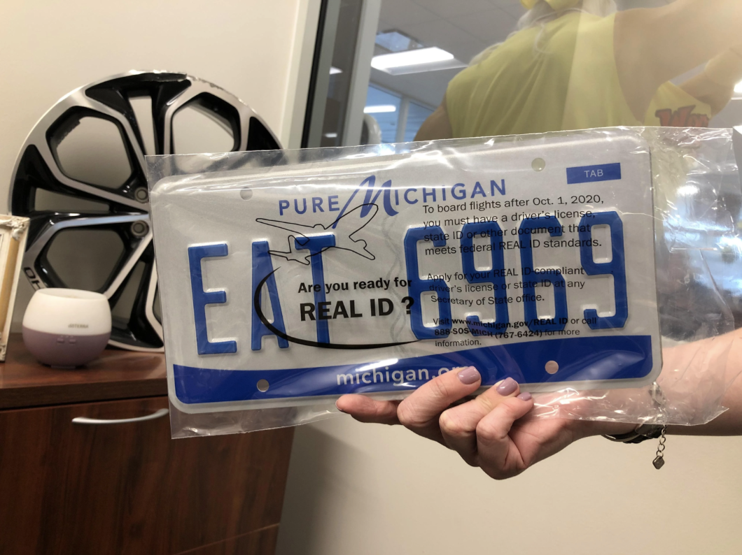Person holding a Pure Michigan license plate that reads &quot;EA7 6193,&quot; alongside a pamphlet. Items on the desk include a wheel rim and a small speaker