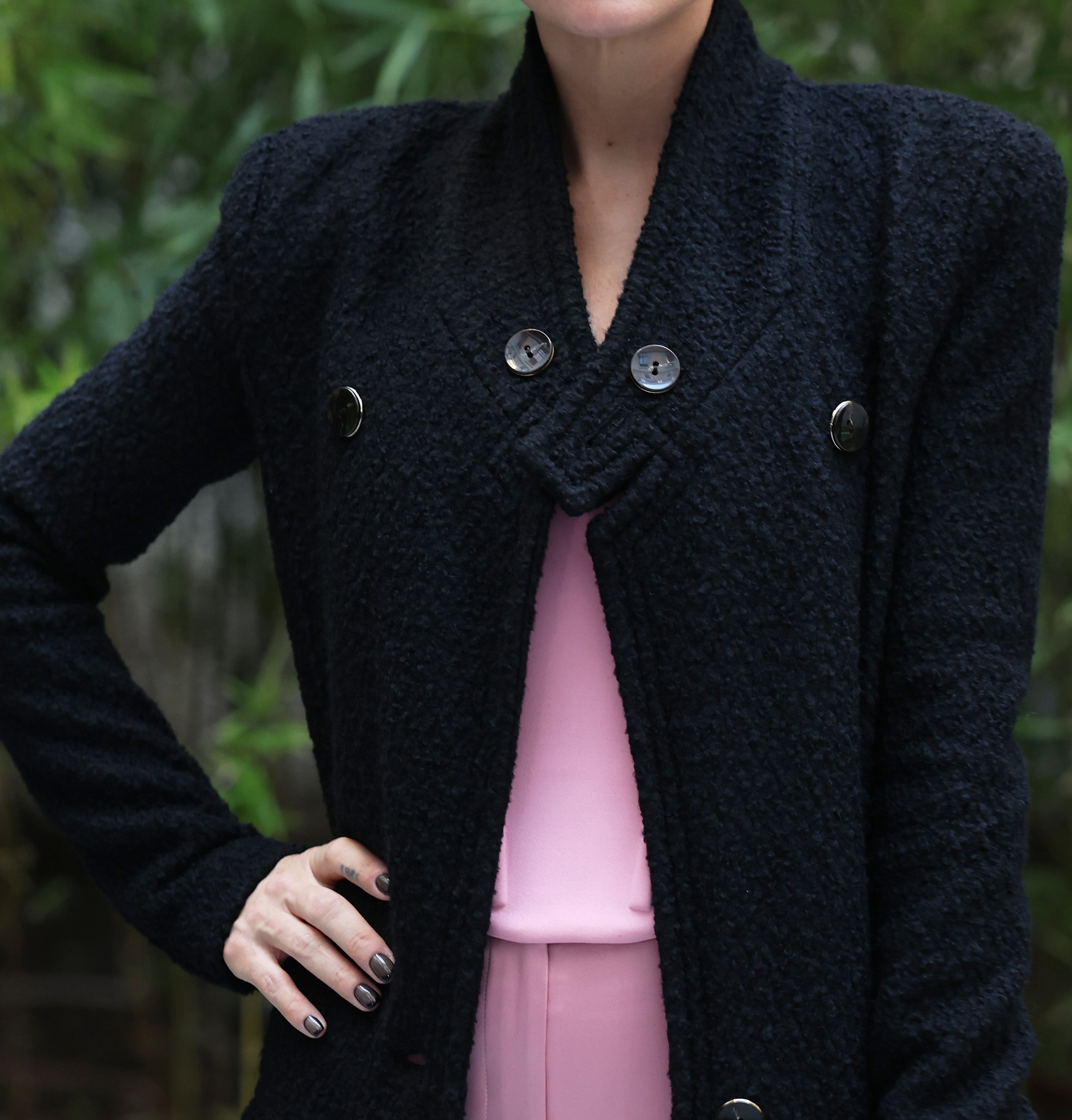 Lily Allen is dressed in a stylish long dark coat over a pink outfit, holding a textured clutch purse. She stands against a backdrop of green foliage