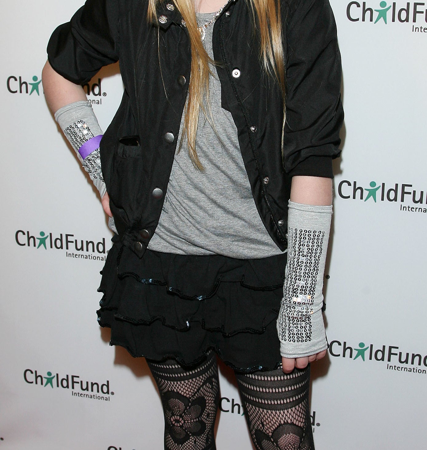Sabrina Carpenter as a kid in a dark jacket, shirt, frilly black skirt, and patterned tights, standing on the red carpet