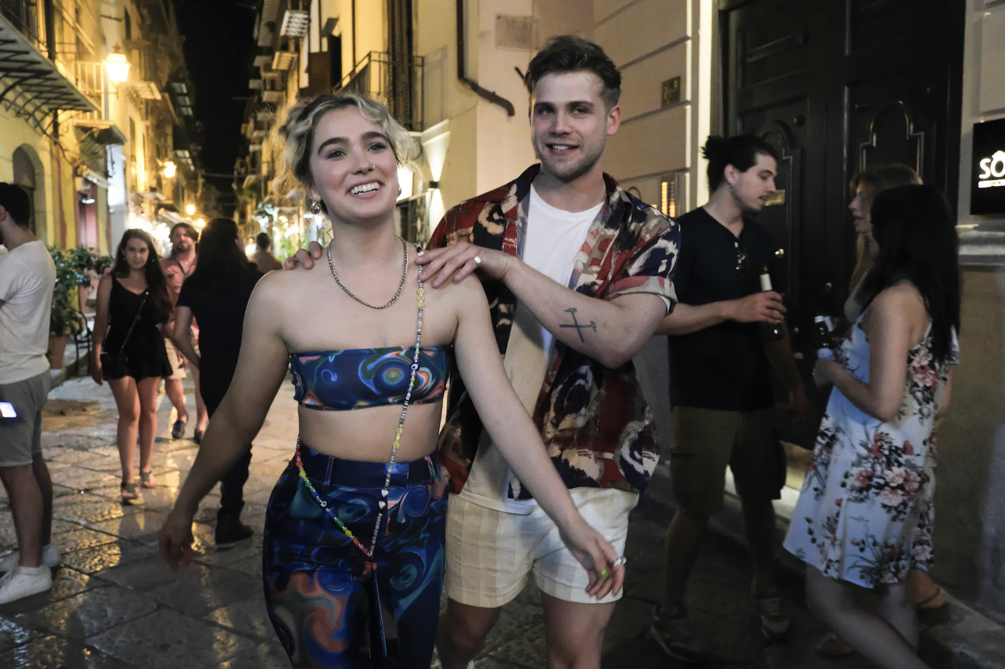 Saoirse Ronan walks with Jack Lowden, who has his hand on her shoulder, on a lively street with people in the background. Ronan wears a patterned crop top and pants