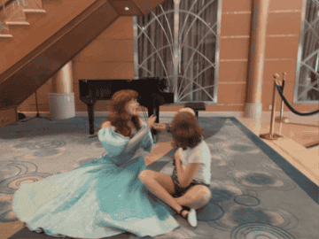 Merida from Disney&#x27;s Brave animated movie sits on the floor next to a young fan, sharing a moment under a staircase indoors