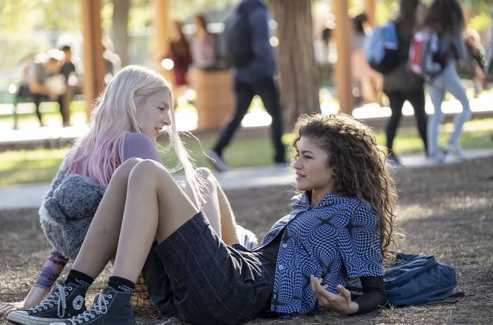 Hunter Schafer and Zendaya sitting on the ground in an outdoor setting, with people walking by in the background