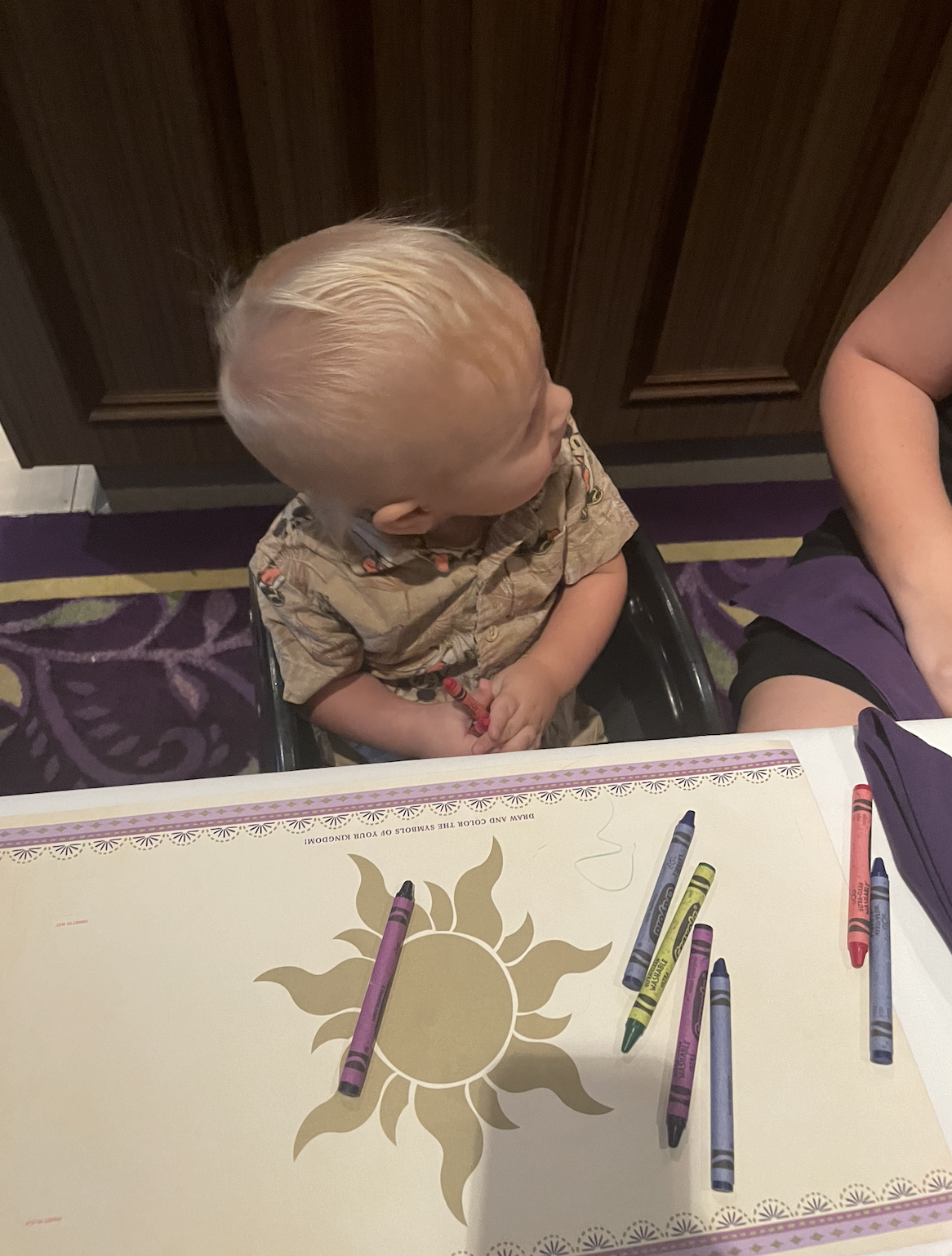 A toddler with light hair sits at a table with crayons and a sun-themed mat, looking to the side. An adult&#x27;s arm is partially visible next to the child