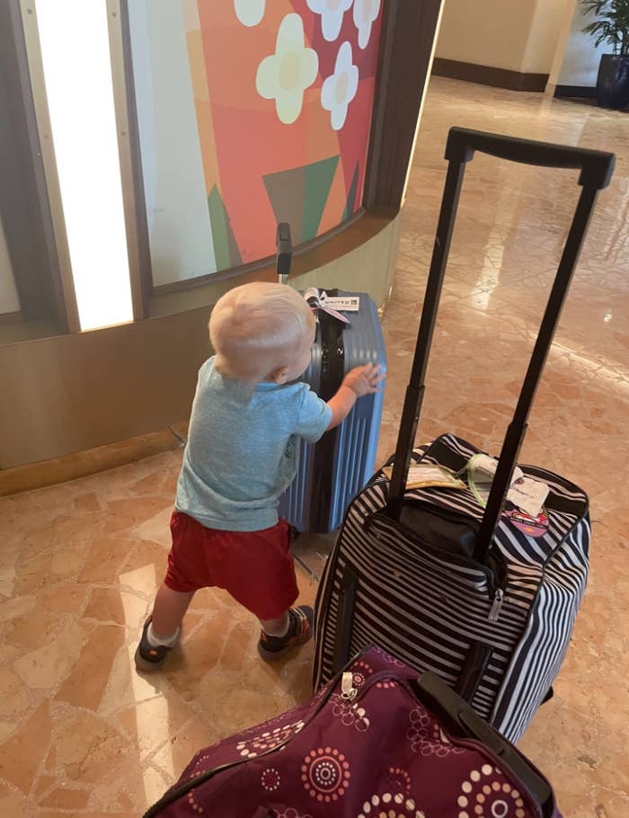 A young child pushes a suitcase in an airport or hotel lobby. Two other suitcases are nearby