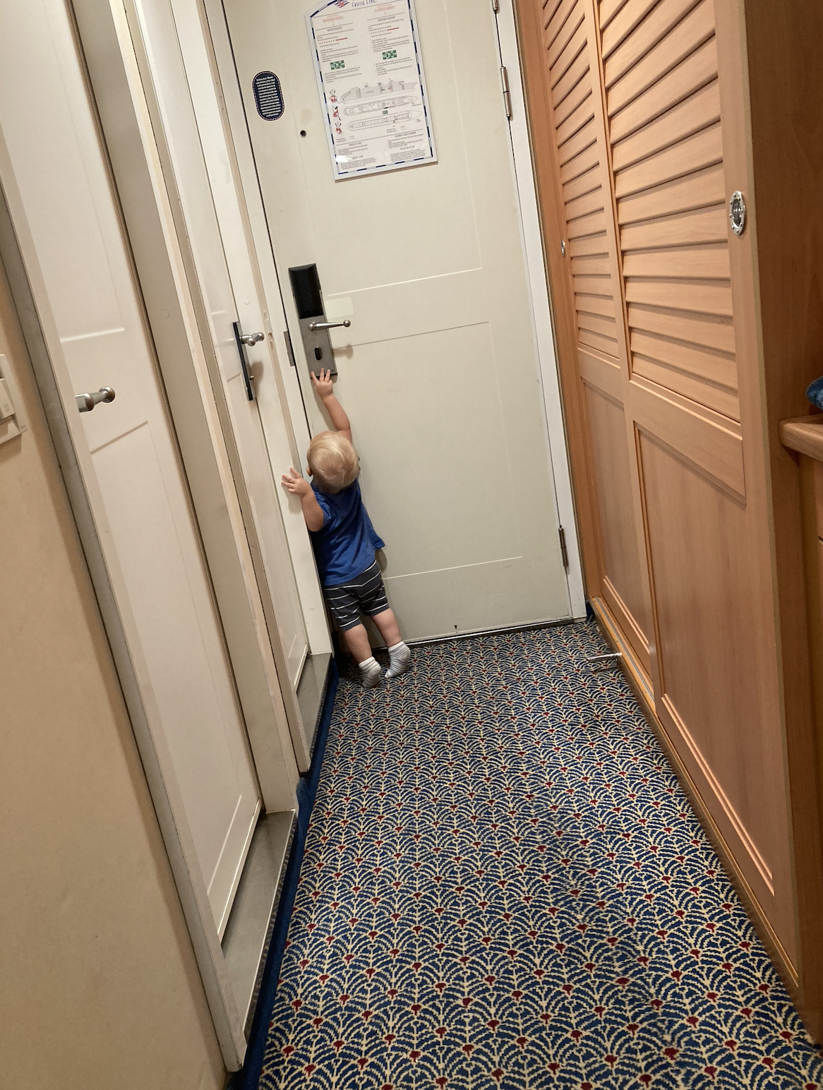 A toddler stands on a patterned carpet, reaching up towards a door handle in an indoor hallway