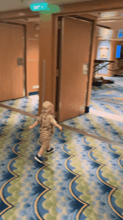 A small child runs playfully down a carpeted hallway on a ship, with interior doors and furniture visible in the background