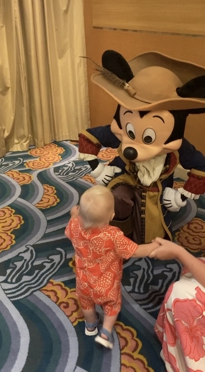 A baby in a patterned outfit approaches Mickey Mouse dressed in a pirate costume while holding an adult&#x27;s hand