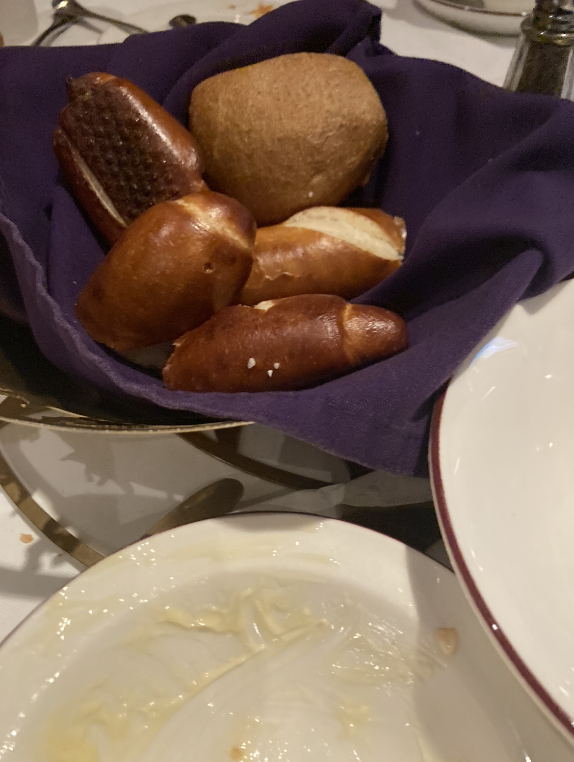 A basket lined with a cloth, filled with assorted bread rolls, sits on a dining table beside an empty dish with remnants of food