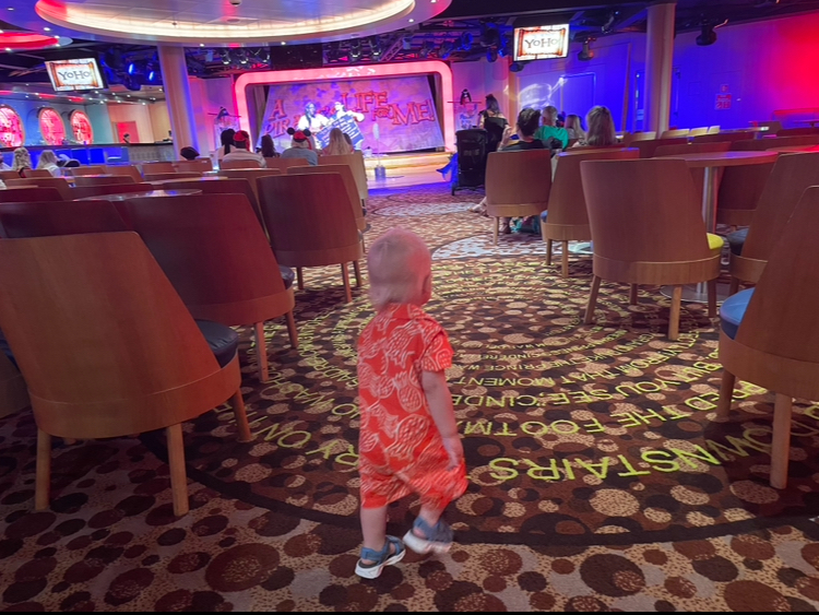 A toddler in patterned clothing walks through an indoor entertainment venue with an illuminated stage in the background. Several people are seated watching a performance