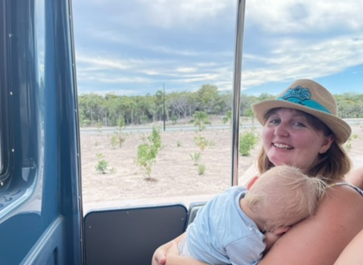 A woman wearing a hat holds a sleeping baby on a bus or train, with a view of a dry landscape and trees outside the window. Names unknown