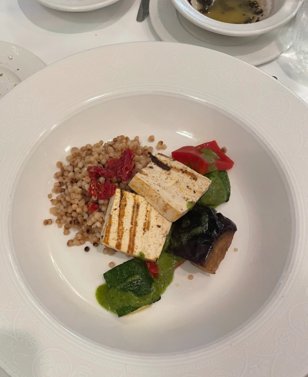 A plated dish featuring grilled tofu, barley, dried tomatoes, and assorted vegetables with a green sauce, served in a white bowl
