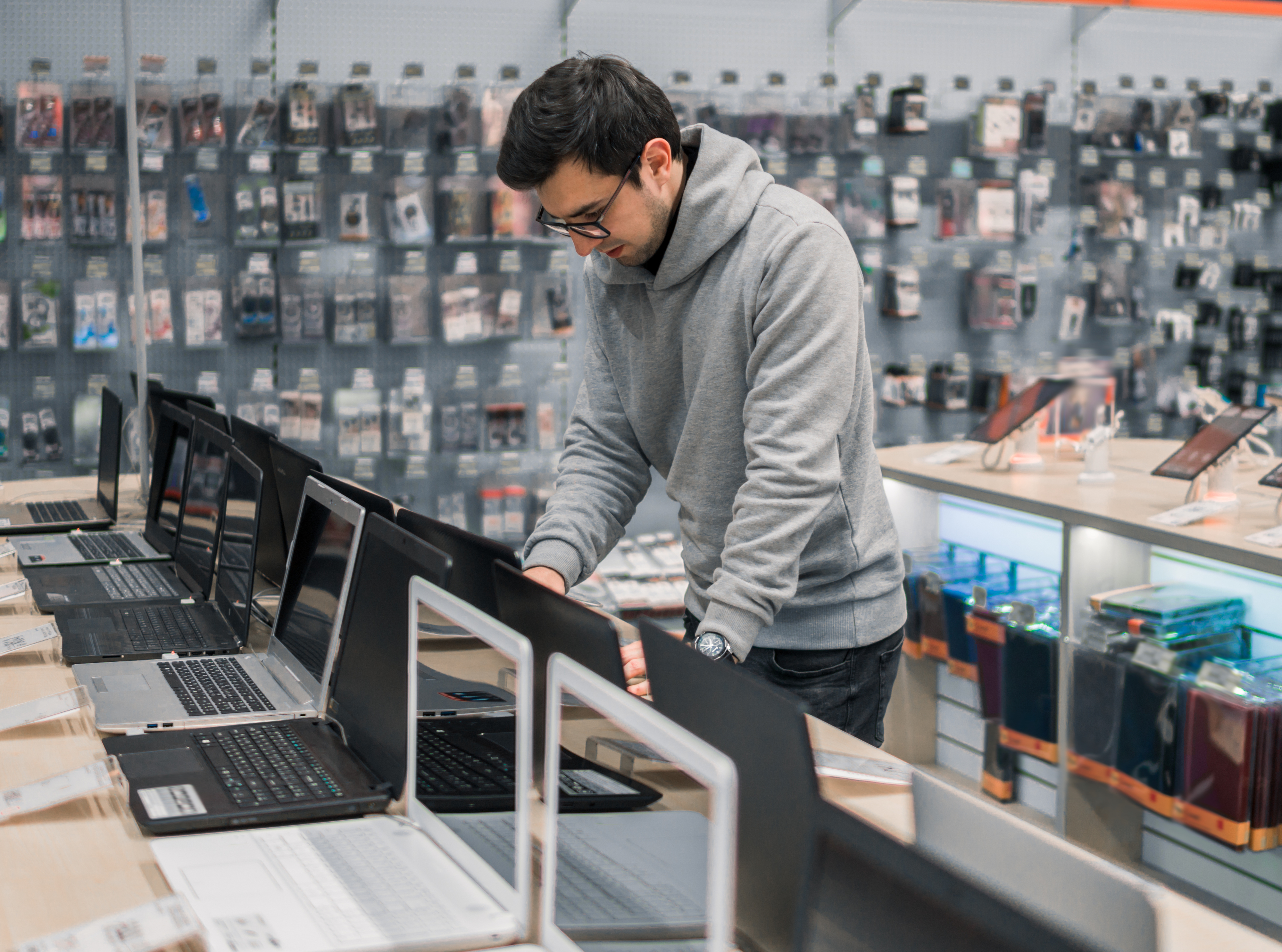 A man wearing a grey hoodie is looking at laptops displayed in a store. The store has many electronic accessories on the shelves