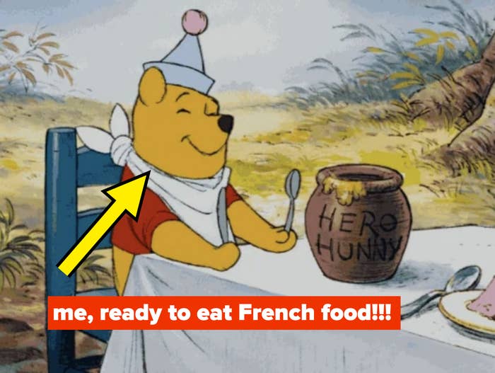 Winnie the Pooh sits at a table with a &quot;HERO HUNNY&quot; pot, wearing a bib and a party hat, happily holding a spoon, ready to eat with the caption: &quot;me, ready to eat French food&quot;