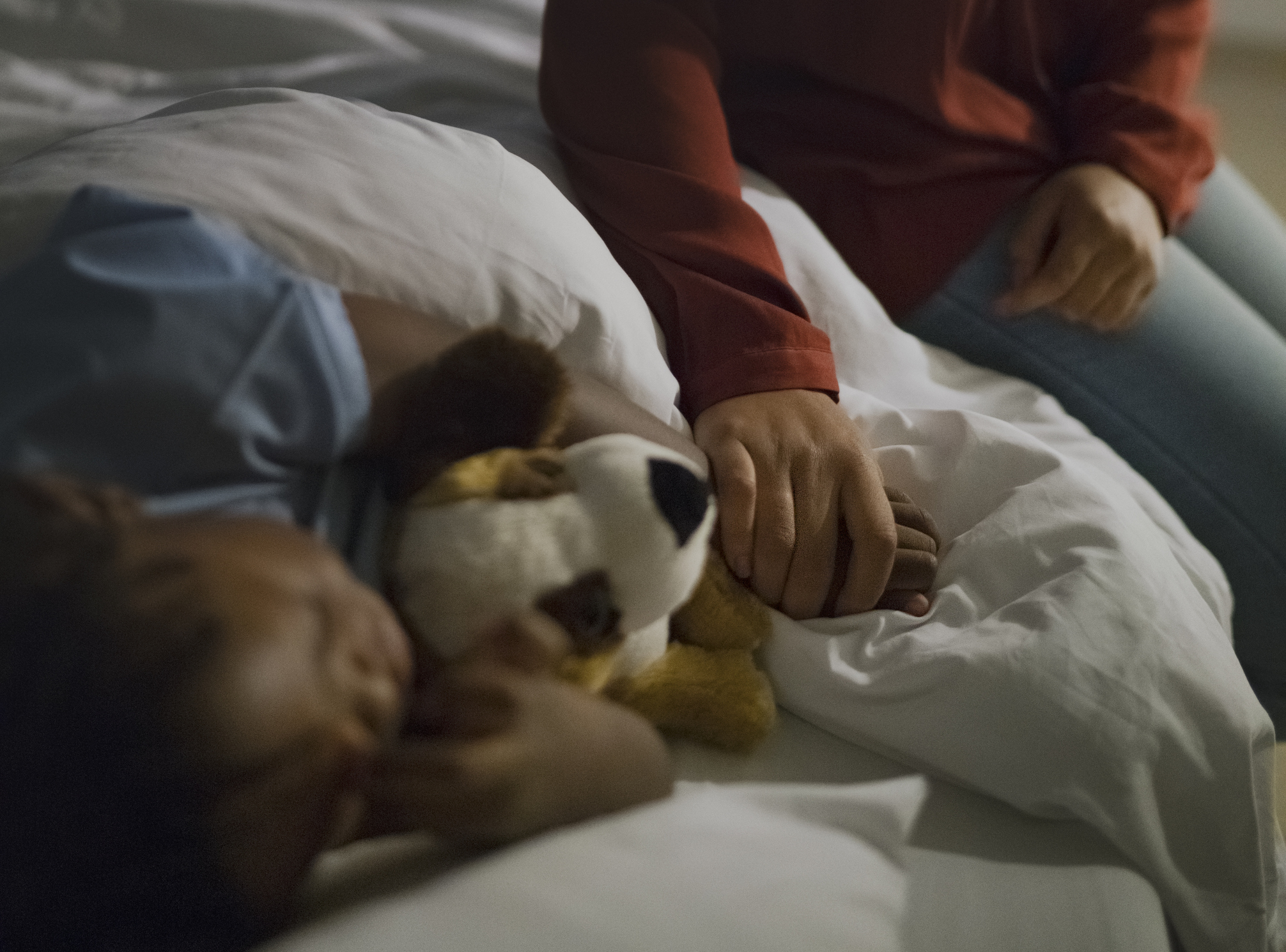 Close-up of a parent holding hands with a child who is lying in bed with a stuffed animal