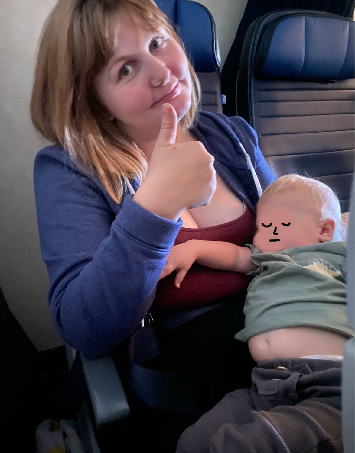 A woman sits on an airplane holding a sleeping baby, giving a thumbs up to the camera