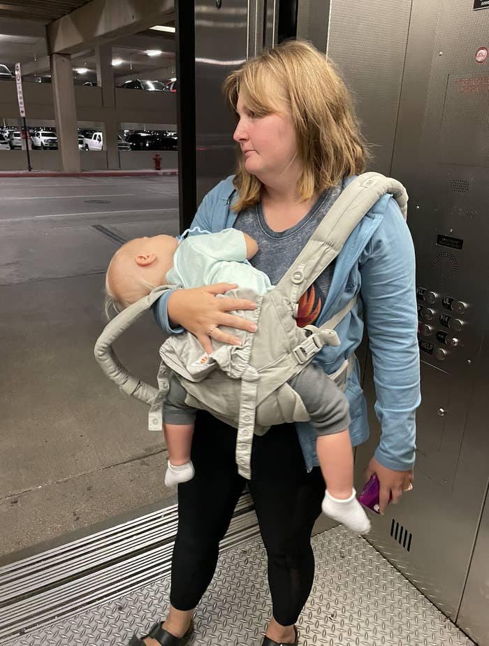 A woman carrying a sleeping baby in a front baby carrier stands inside an elevator