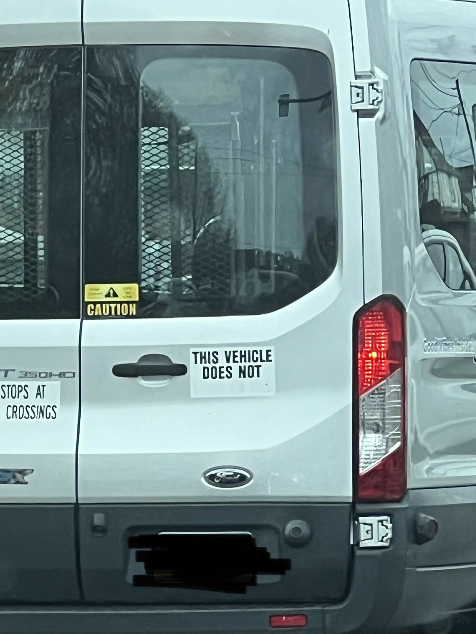 Back of a white van featuring a caution sign, text reading &quot;This vehicle does not stop at crossings,&quot; and a black stripe concealing a part of the license plate
