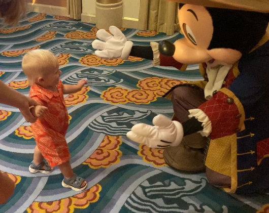 A baby, held by an adult, walks towards Mickey Mouse who is kneeling with arms open. Mickey is dressed in a colorful costume with long sleeves and gloves