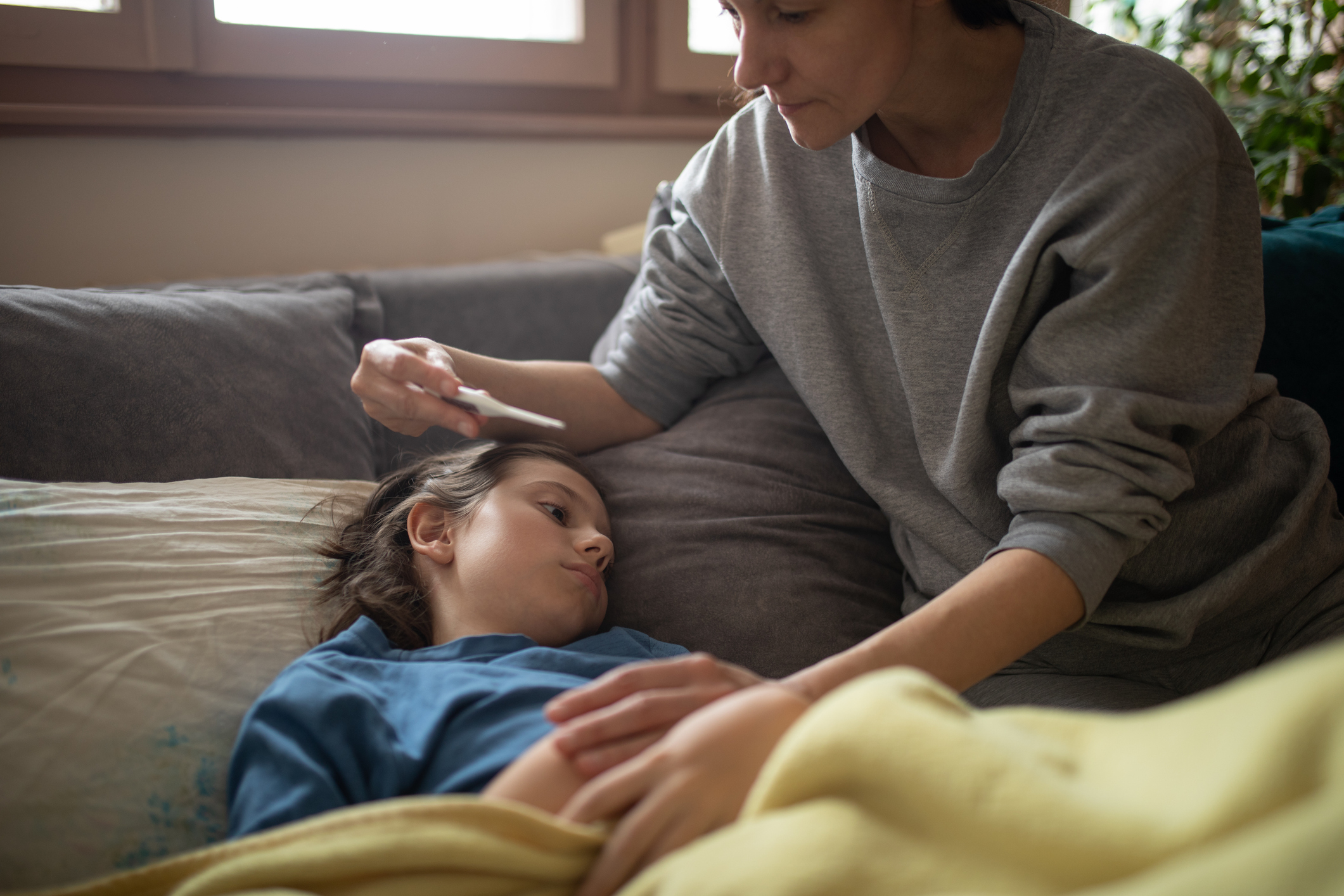 A woman in a gray sweatshirt is seated on a couch, placing a thermometer in the mouth of a resting child covered by a yellow blanket