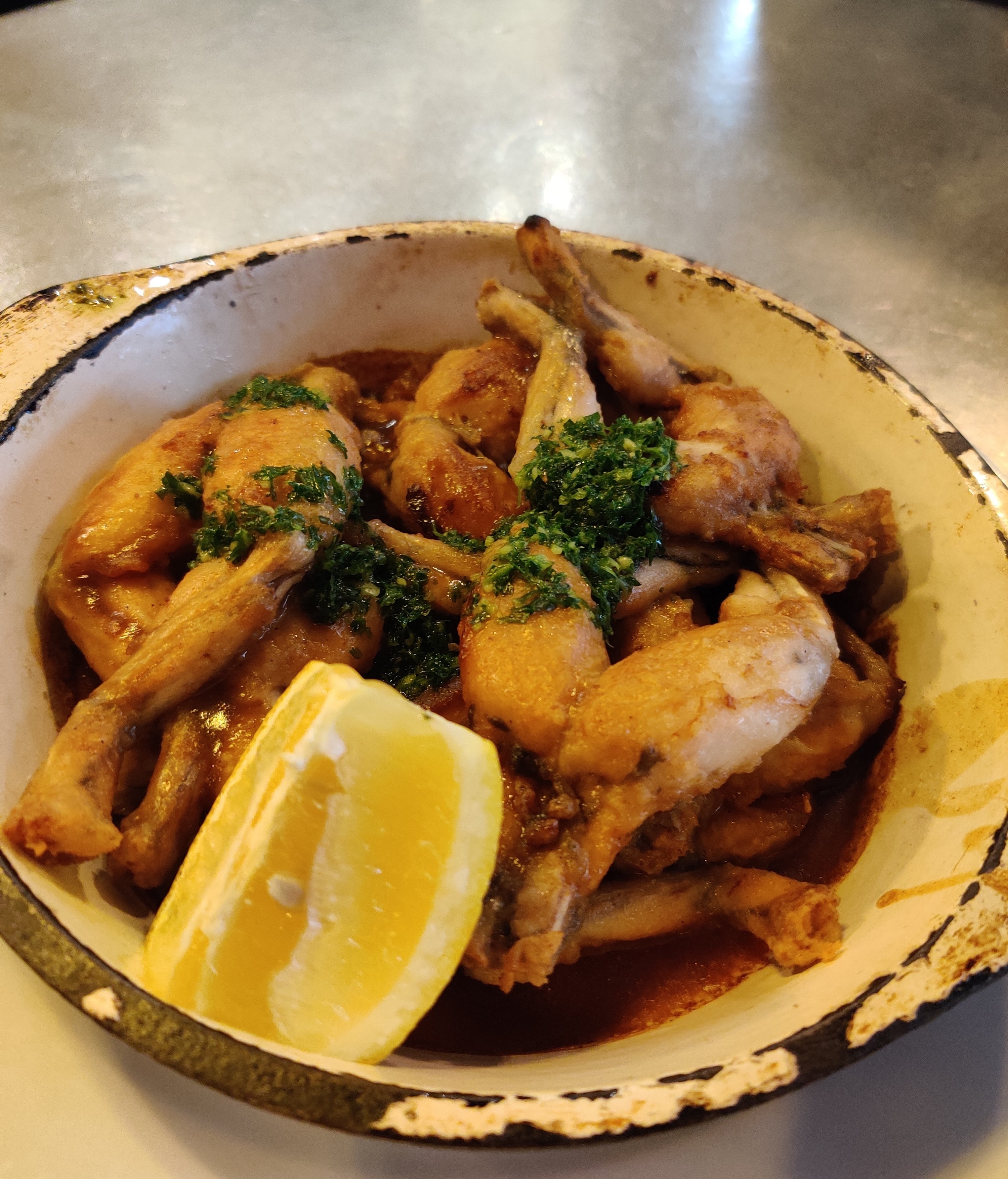 A rustic dish of frog legs garnished with green herbs and served with a lemon wedge