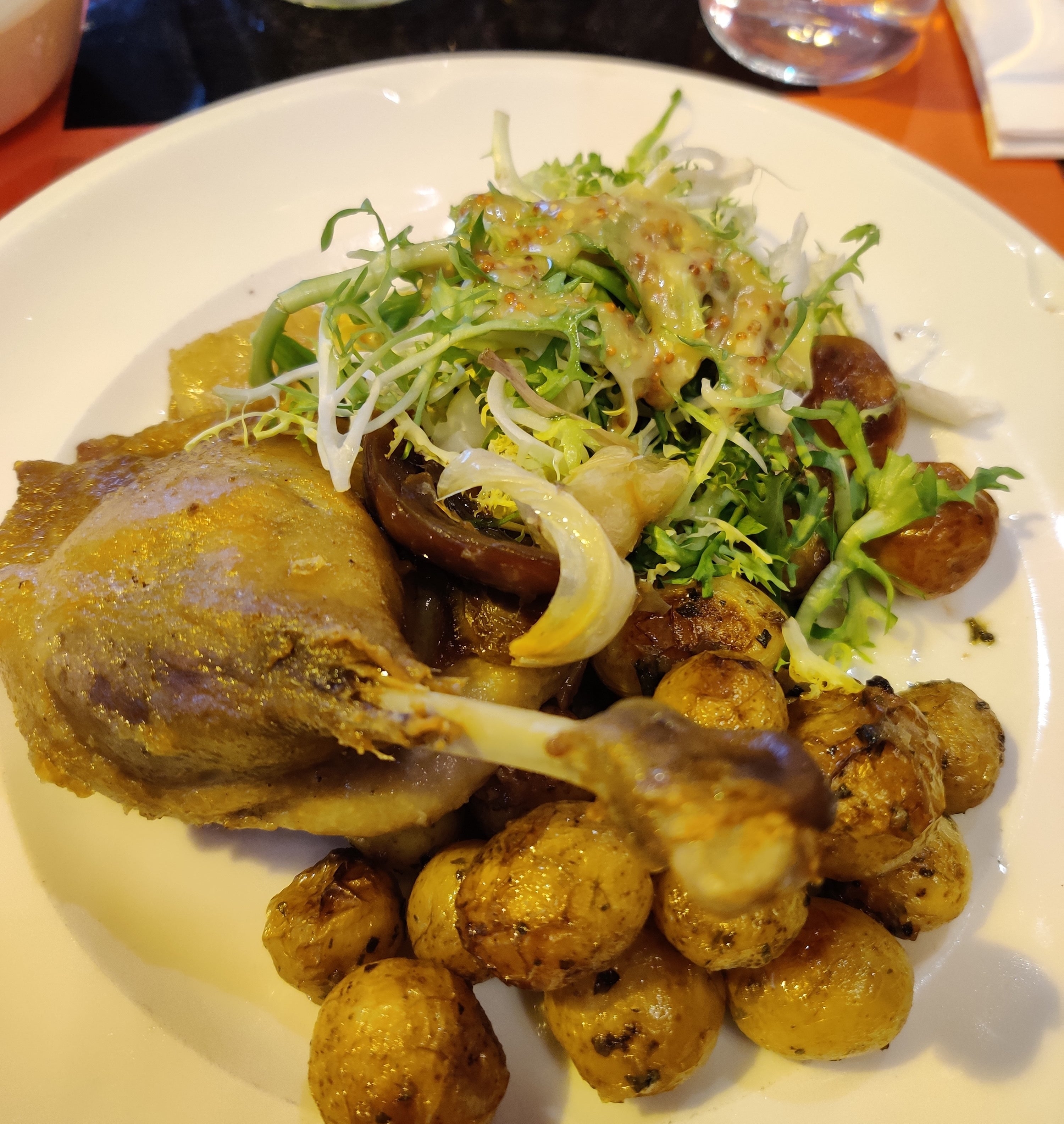 A plate of duck confit served with crispy baby potatoes, frisée salad, grilled mushrooms, and drizzled with a mustard dressing