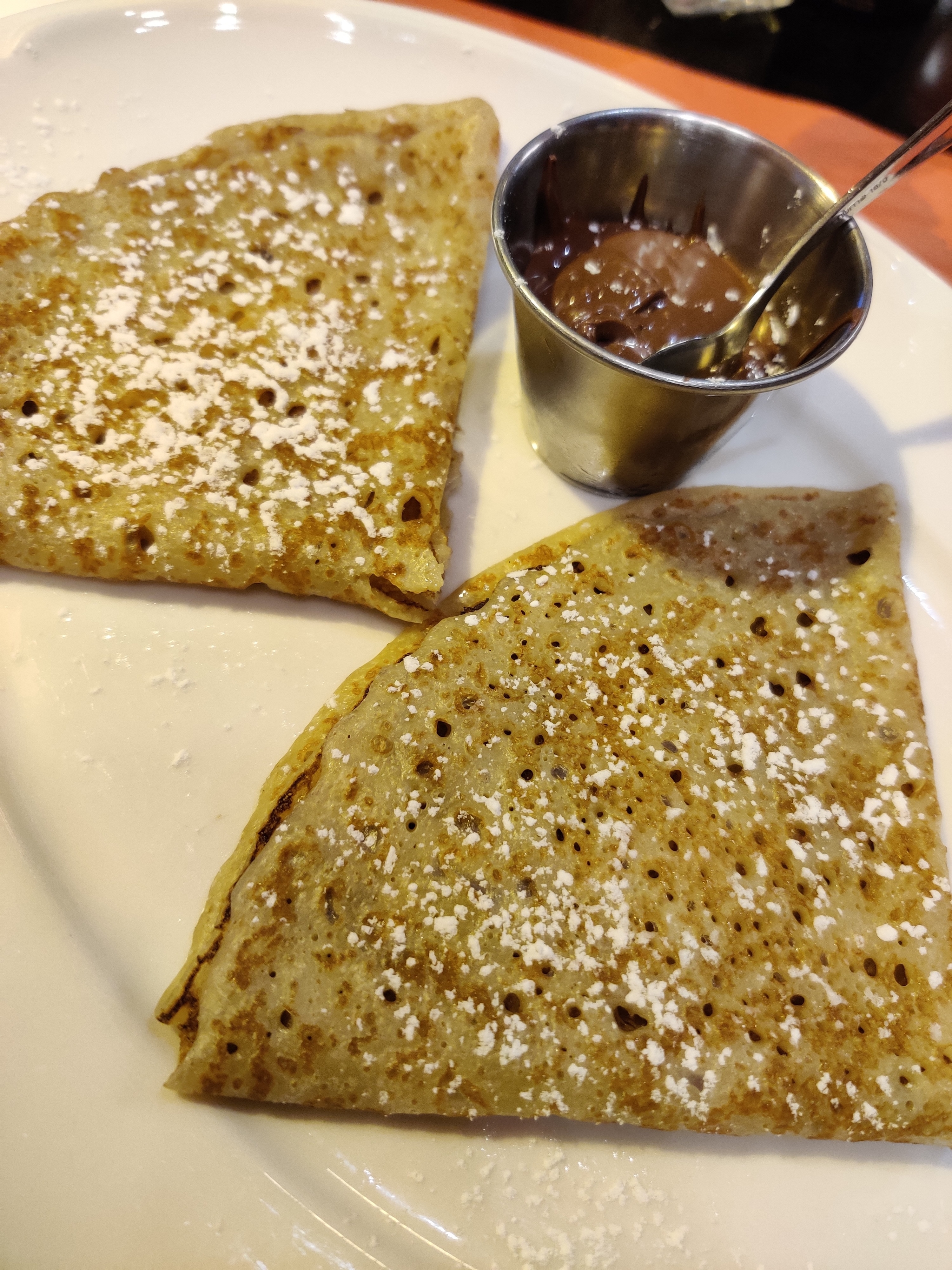 Two triangular crepes lightly dusted with powdered sugar, served with a small metal cup of nutty chocolate spread