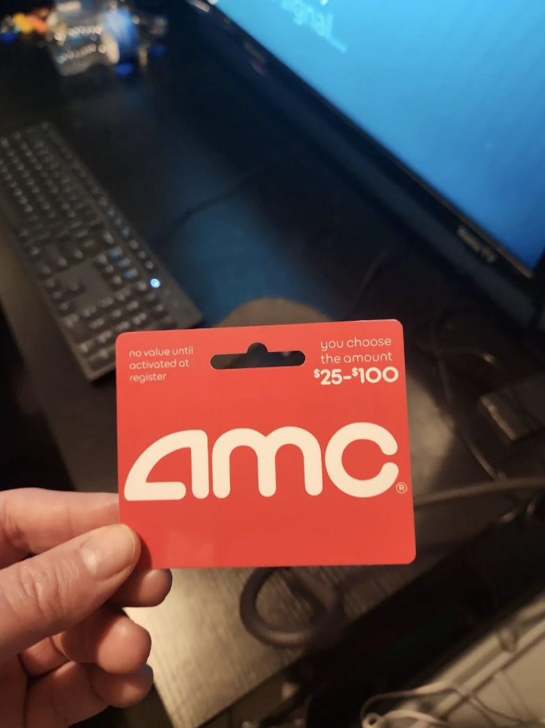 Hand holding an AMC gift card valued between $25-$100, in front of a computer setup