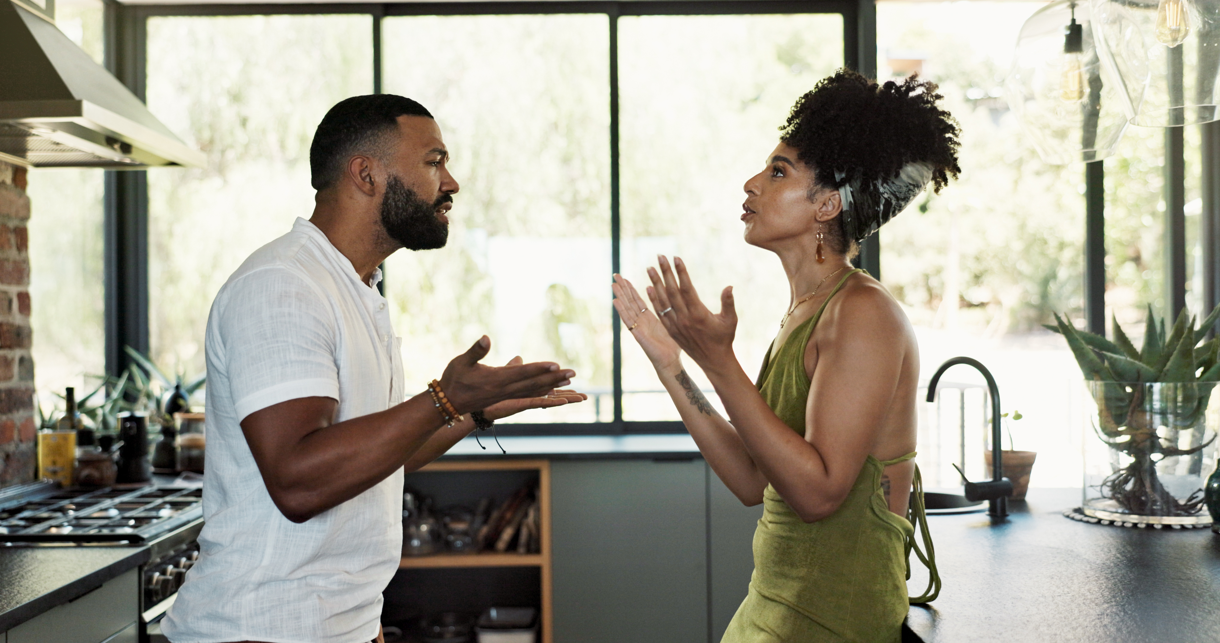 A man and a woman stand in a kitchen, engaged in an intense conversation. The woman wears a sleeveless dress and has her hair up; the man wears a casual button-up shirt