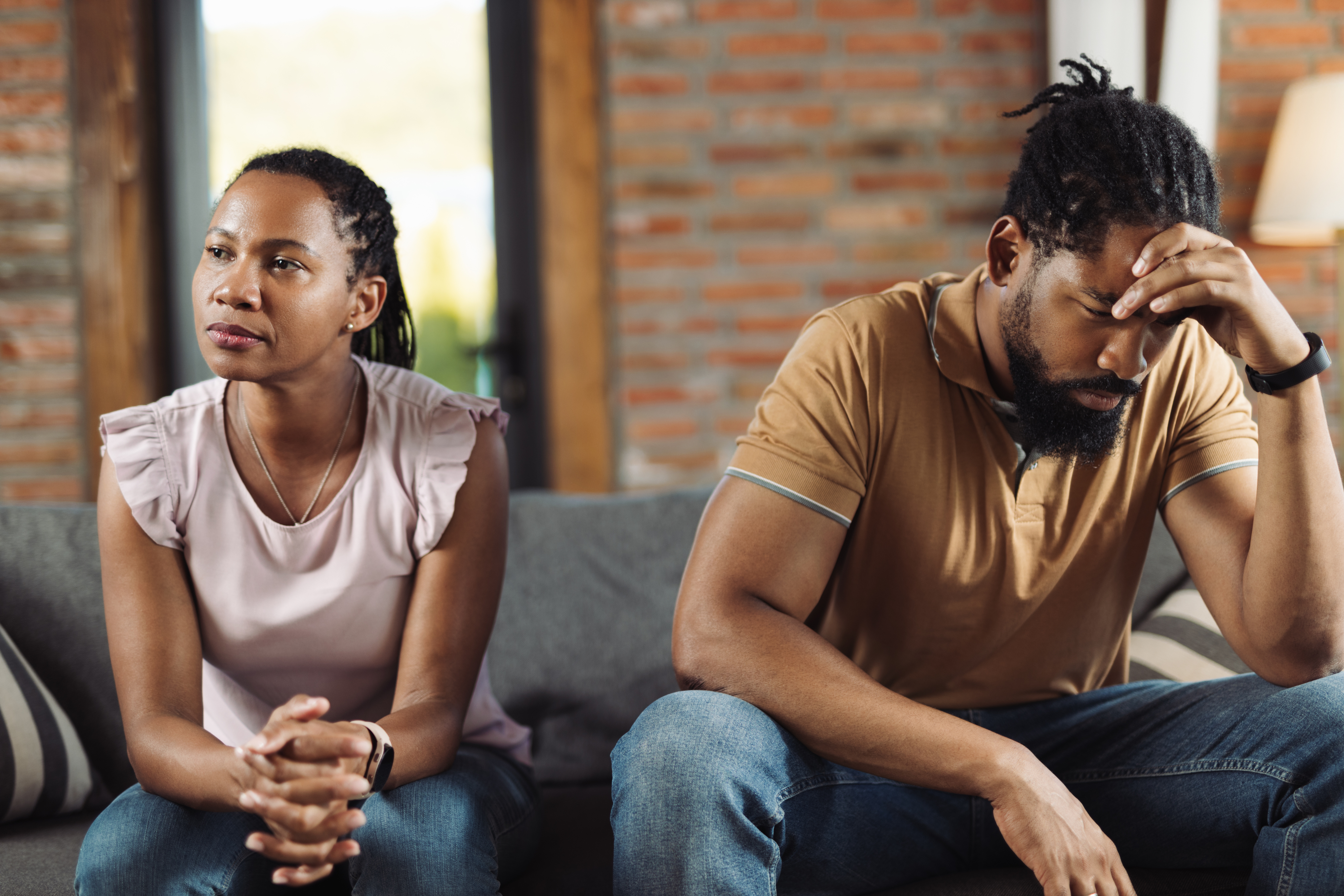 Two people sit on a couch, looking frustrated. The woman has her hands clasped, while the man holds his head in his hand