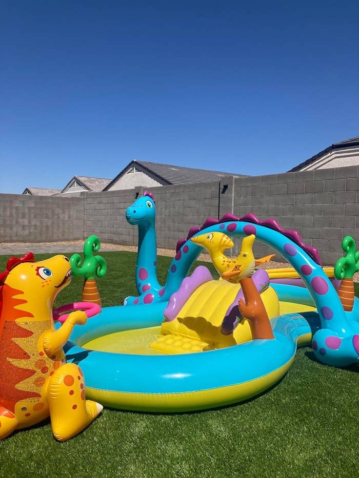 a reviewer photo of the inflatable dinosaur-themed kiddie pool with slides and water sprayers set up in a backyard