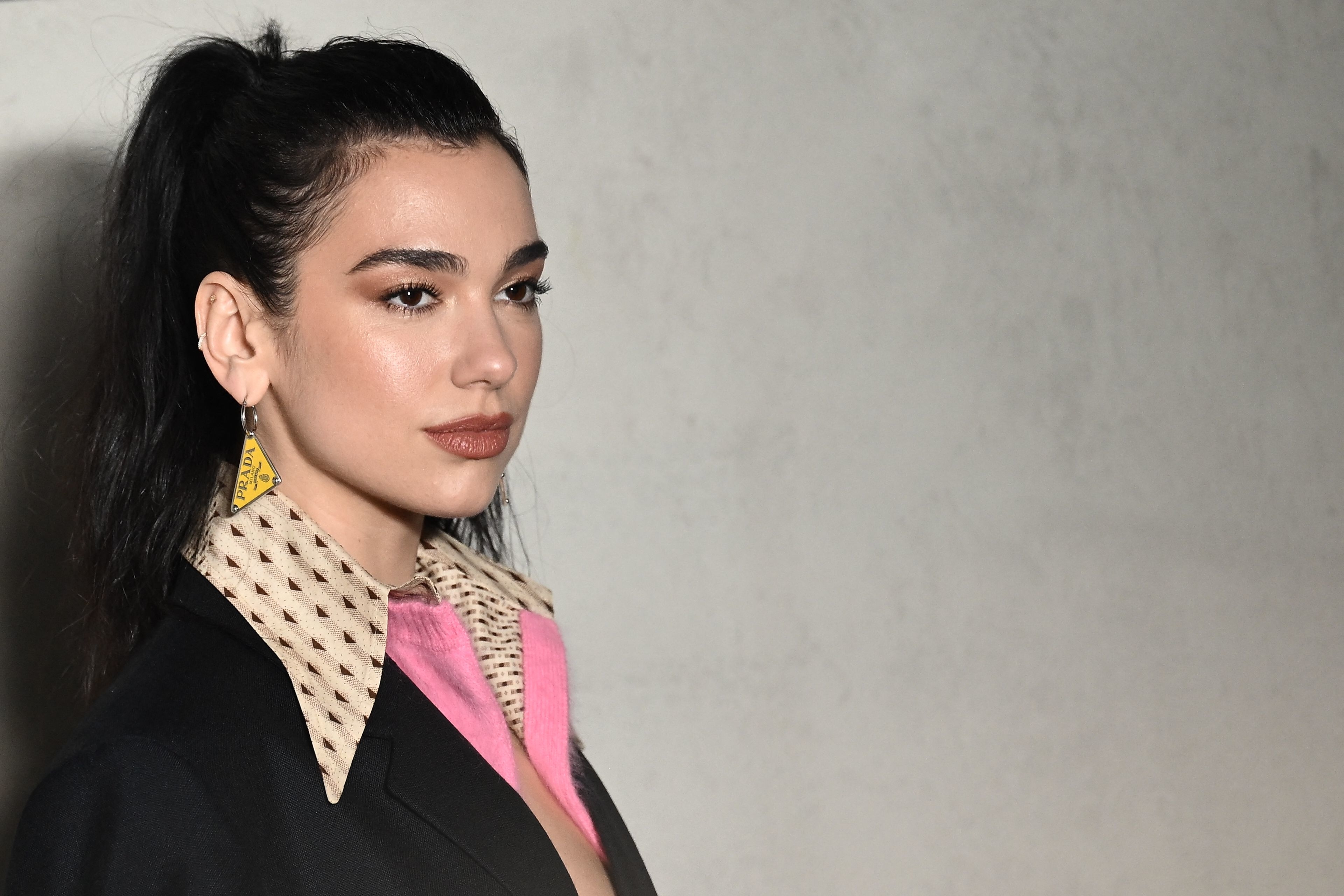 Dua Lipa wearing a stylish, patterned shirt beneath a dark blazer, posing for a photo, with her hair in a high ponytail