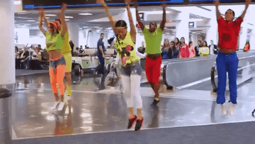 People dancing energetically at an indoor public space with an audience watching. Four dancers wear bright casual clothes