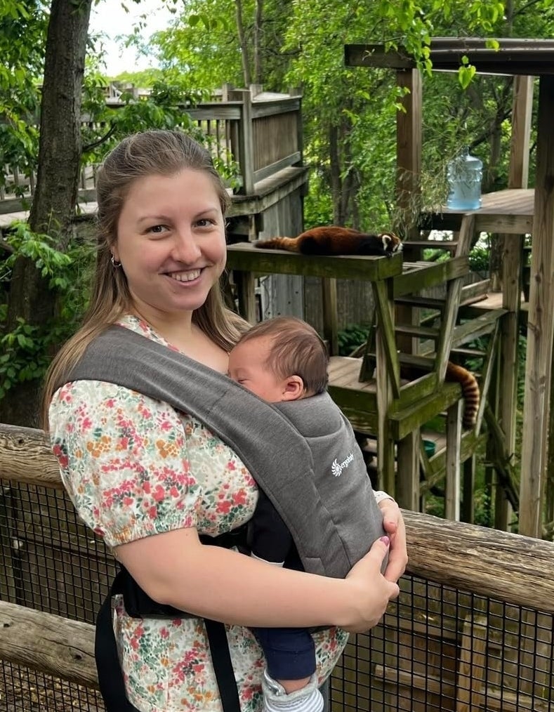 a reviewer wearing a floral blouse smiles while holding a baby in a front carrier with red pandas in the background