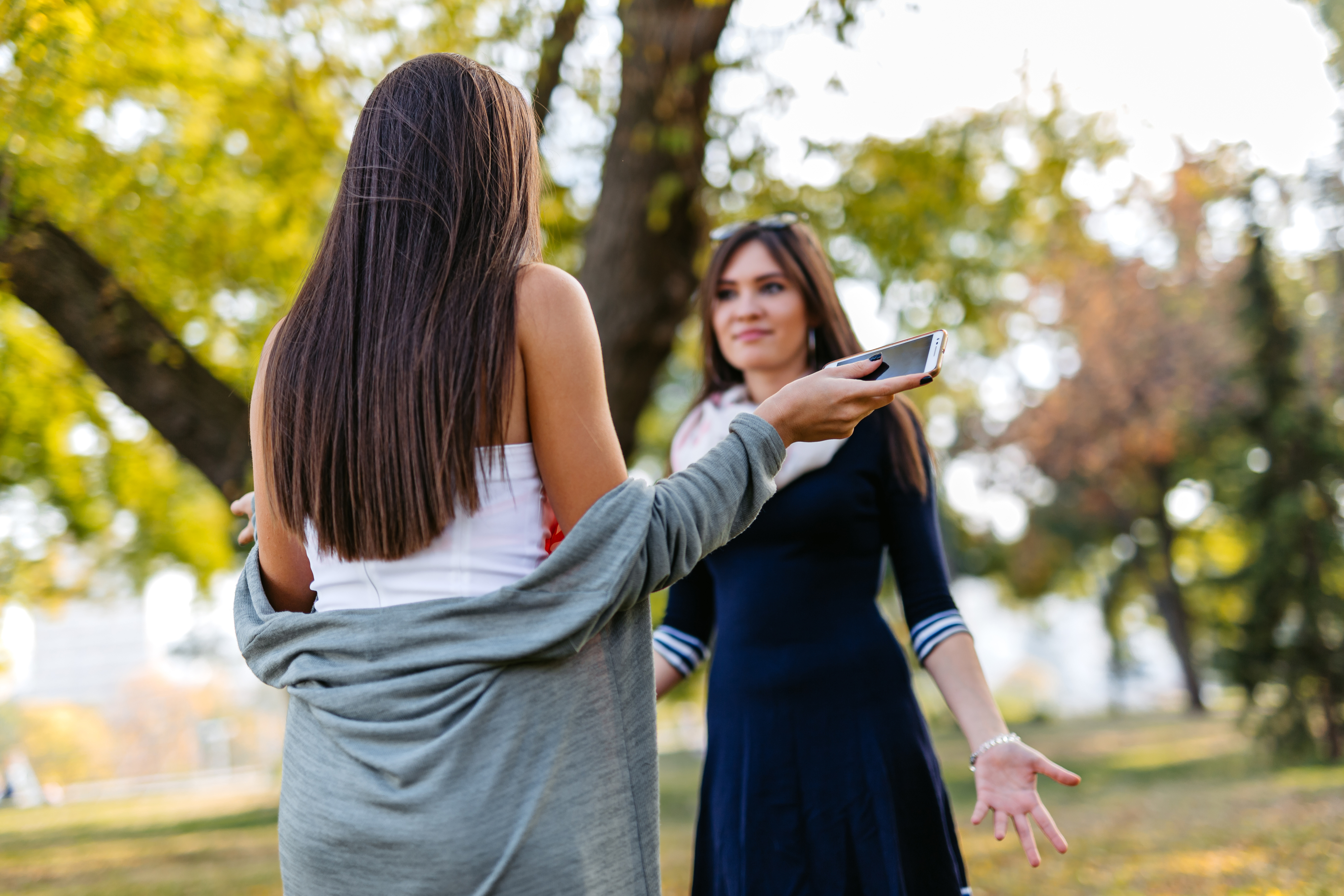 Two women talking outdoors; one holds a smartphone while the other wears a cardigan over a dress. Names of the individuals are not known