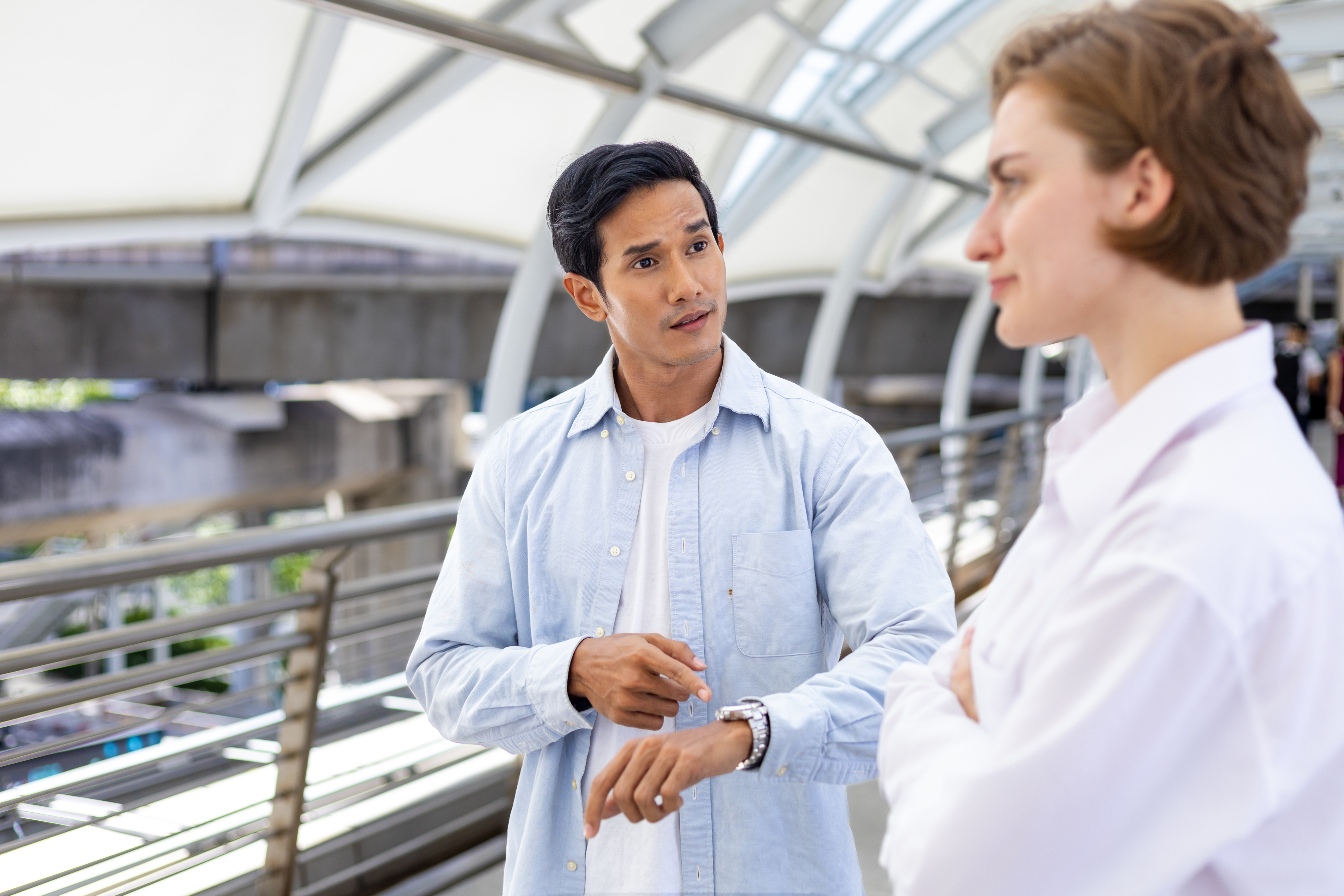 A man and a woman talk on a modern pedestrian bridge. The man points to his watch while looking at her. Both are casually dressed