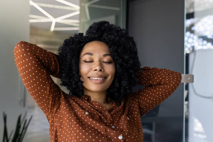 Woman with black curly hair, smiling with eyes closed, in a brown polka dot shirt, relaxing with hands behind her head in an indoor setting