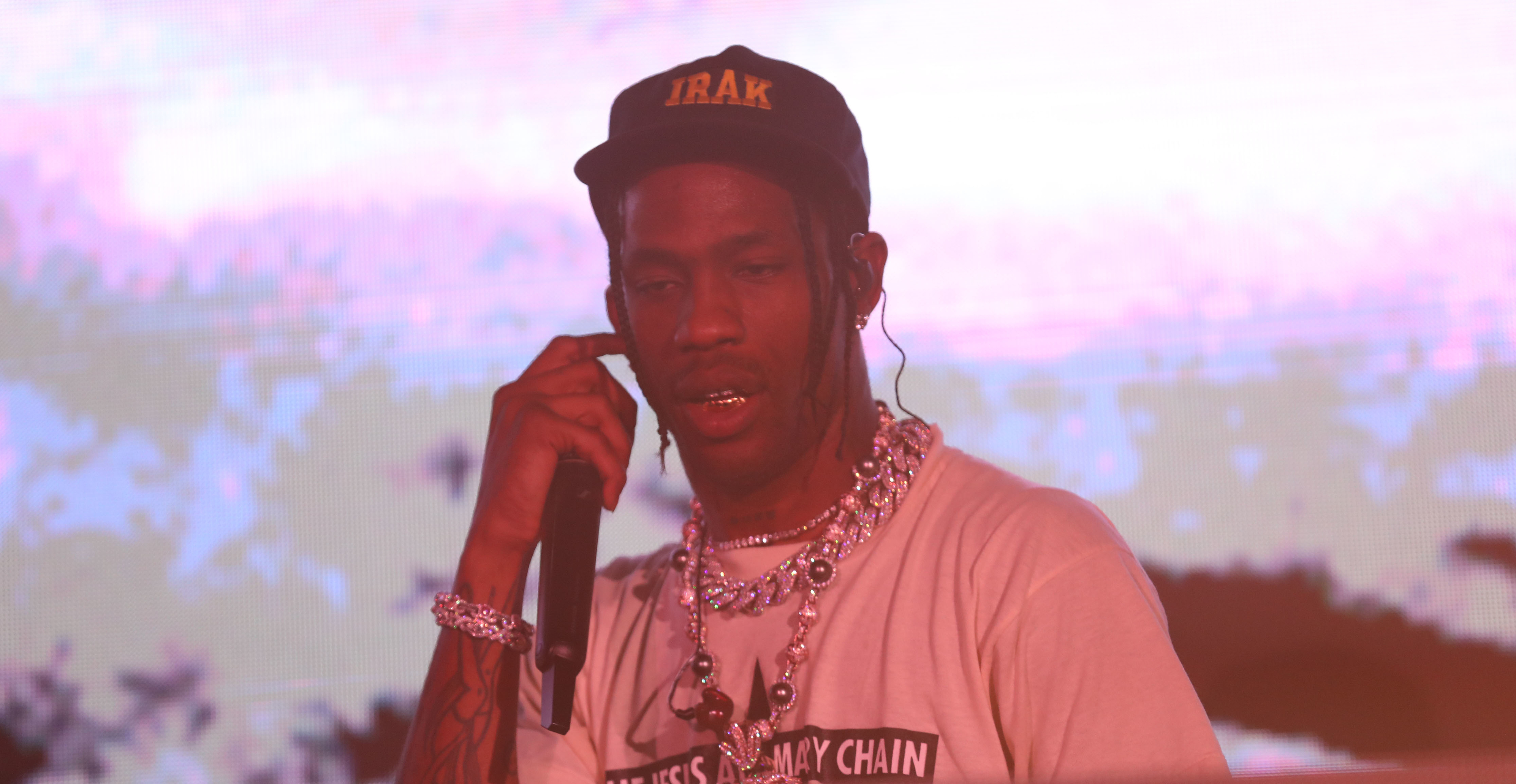 Travis Scott performing on stage, wearing a black cap, white T-shirt, and several silver chains around his neck. He is holding a microphone close to his mouth