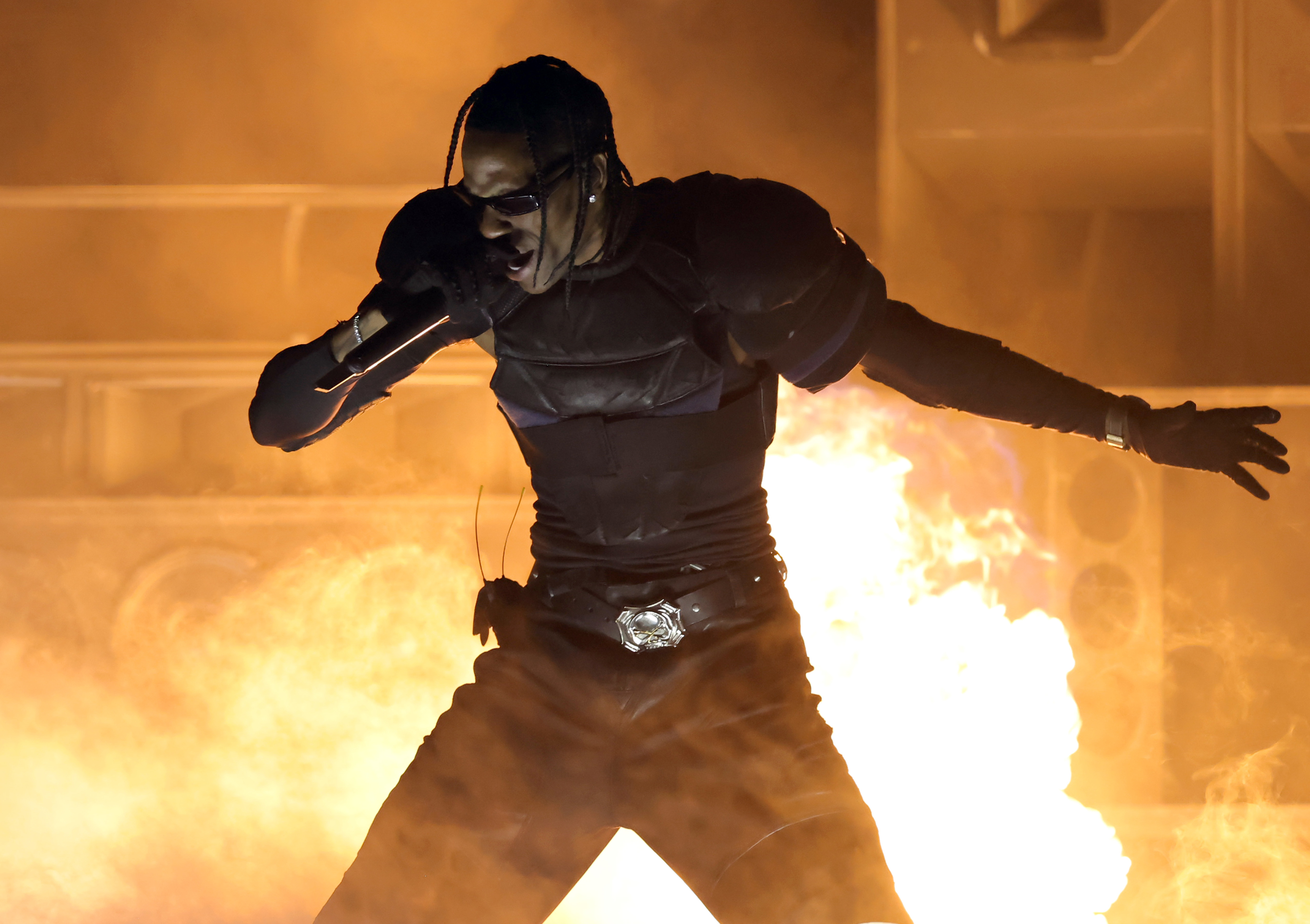 Travis Scott performing energetically on stage, dressed in a futuristic outfit with flames in the background