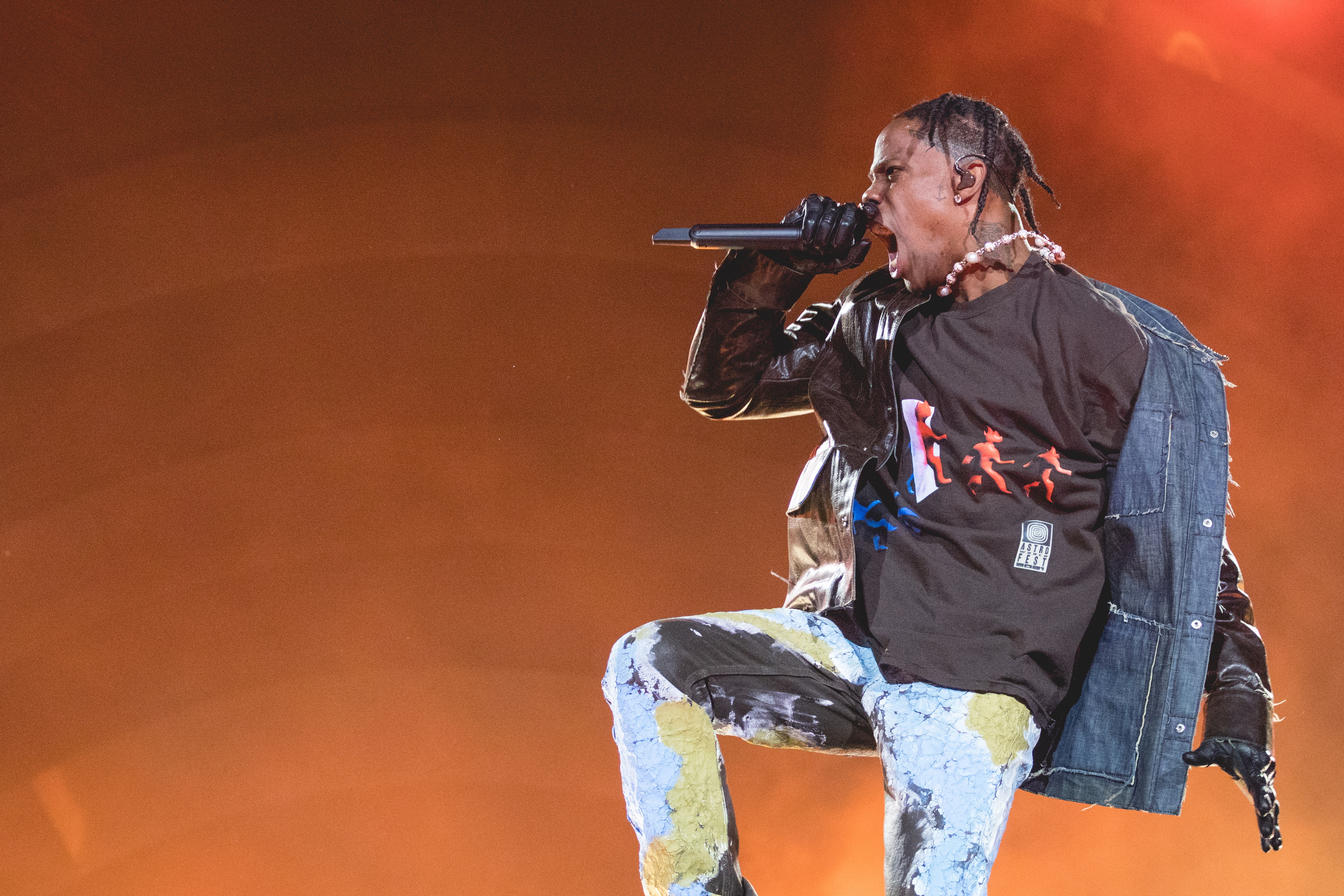 Travis Scott performs energetically on stage, wearing a graphic t-shirt, denim jacket, and distressed jeans while holding a microphone