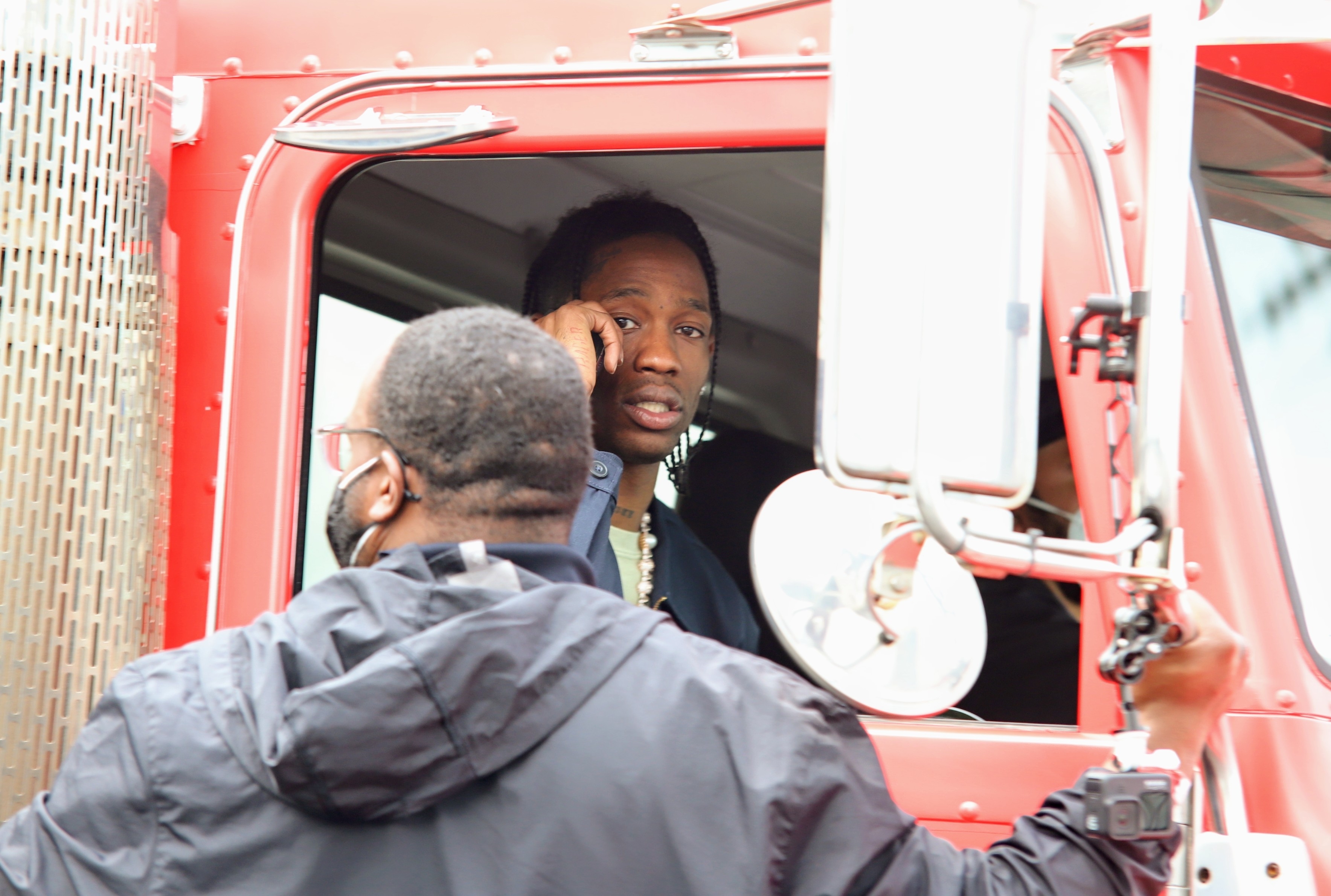 Travis Scott sits in the driver&#x27;s seat of a truck while another man wearing glasses and a jacket speaks to him through the window