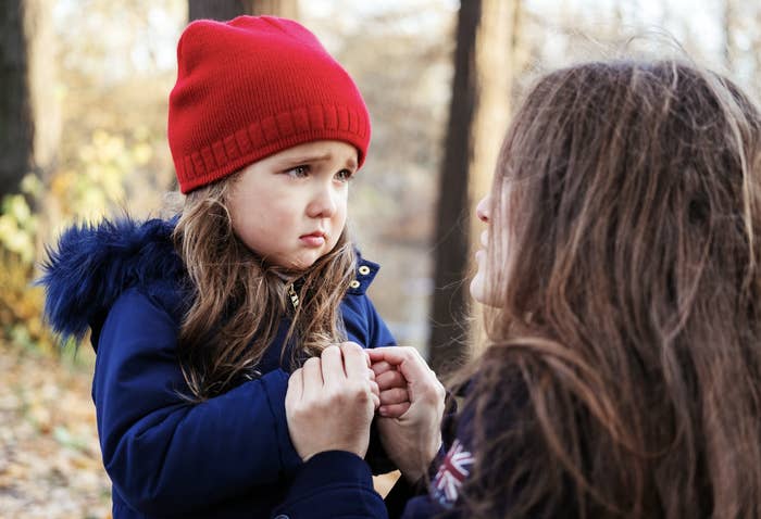 A young girl wearing a red knit hat and blue coat looks sad as she holds hands with a woman, possibly her mother, in a forested area