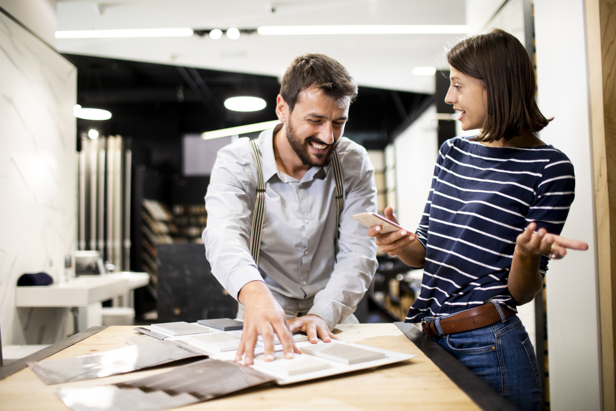 A man and a woman are standing at a table with open material samples. The man is smiling and leaning over to examine the samples while the woman shows him something on her phone