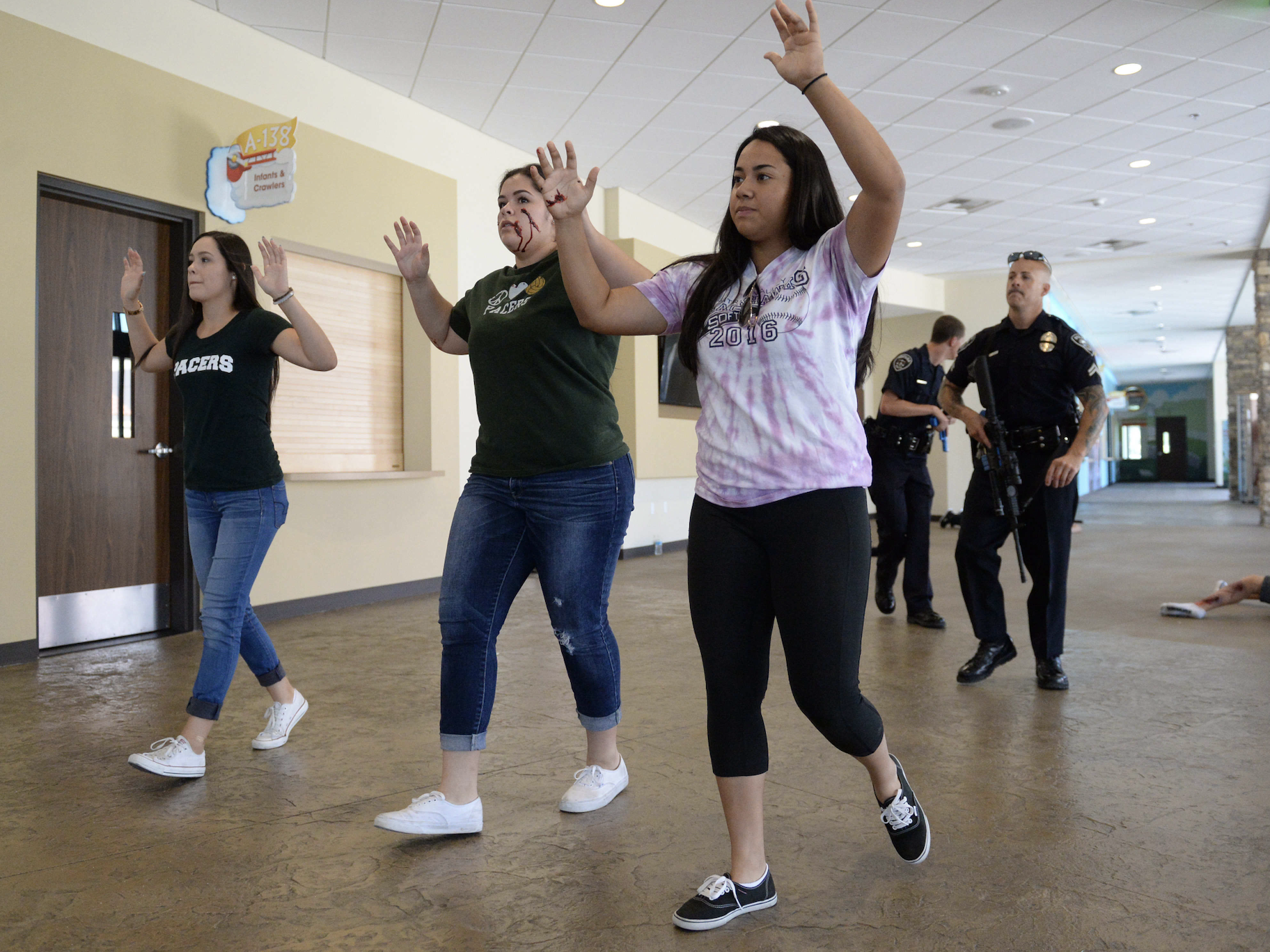 Three people walk with their hands up, followed by two police officers in a school hallway