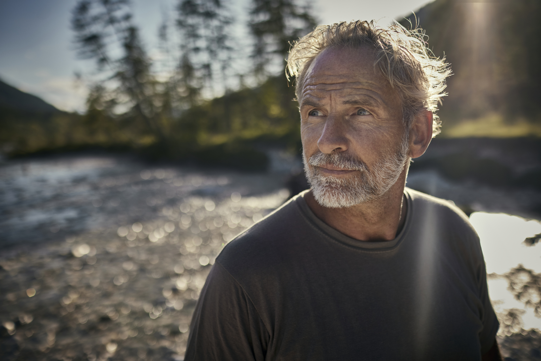 Older man with gray hair and a beard looks thoughtful at a scenic outdoor setting with trees and water in the background