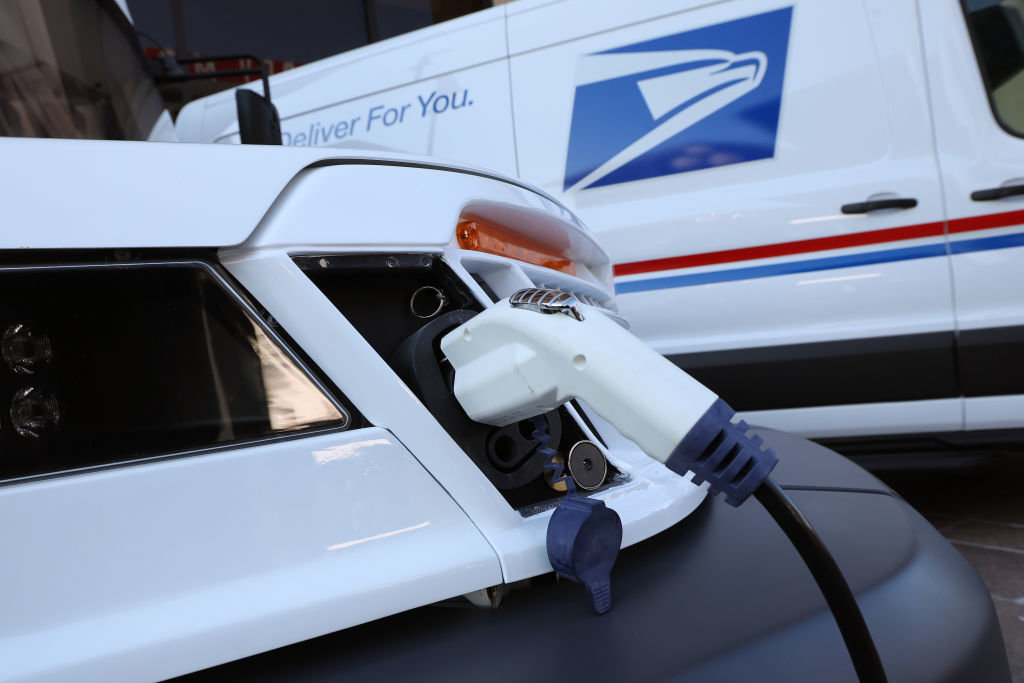 An electric vehicle is being charged with a postal service van in the background. The van bears the U.S. Postal Service logo and slogan &quot;Deliver for You.&quot;