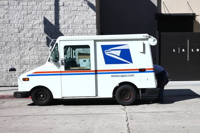 USPS mail truck parked on a street near a building with a person partially visible at the rear door