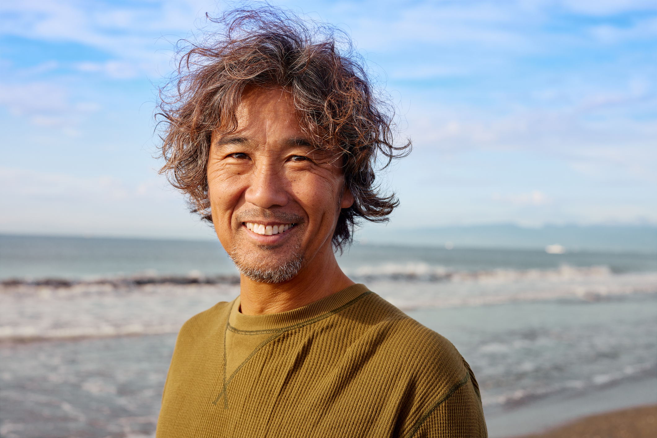 A person with wavy hair and a smile stands on a beach with the sea and sky in the background