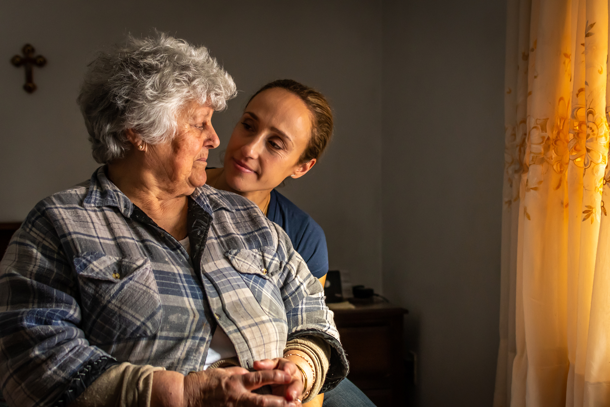 An elderly woman with a plaid shirt gazes at a younger woman in a blue top in a dimly lit room, conveying a moment of connection and care