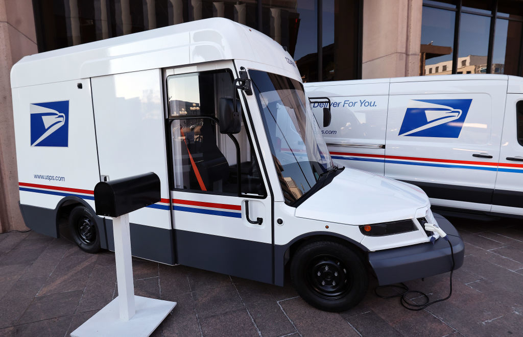 A new USPS electric mail delivery vehicle is parked next to a mailbox and another USPS van outside a building